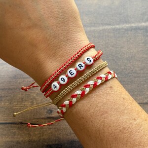May include: Four handmade bracelets in red, white, and tan. One bracelet spells out "49ERS" with white letter beads. Other bracelets feature braided and woven designs. The bracelets are on a person's wrist.