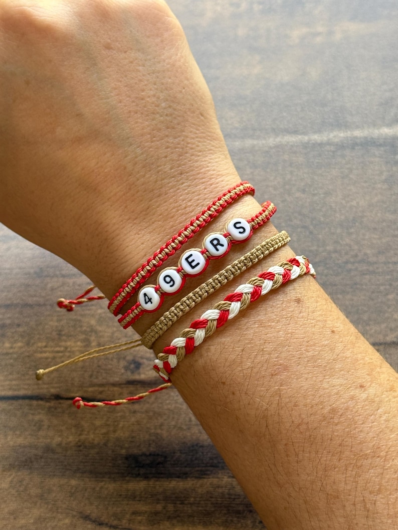 May include: Four handmade bracelets in red, white, and tan. One bracelet spells out "49ERS" with white letter beads. Other bracelets feature braided and woven designs. The bracelets are on a person's wrist.
