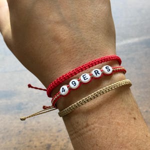 May include: Three bracelets stacked on a wrist. The top and middle bracelets are red, with the middle one spelling "49ERS" with white letter beads. The bottom bracelet is tan.