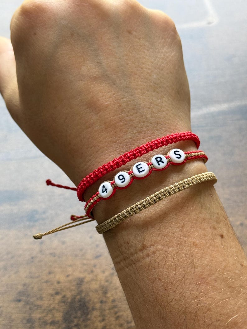 May include: Three bracelets stacked on a wrist. The top and middle bracelets are red, with the middle one spelling "49ERS" with white letter beads. The bottom bracelet is tan.