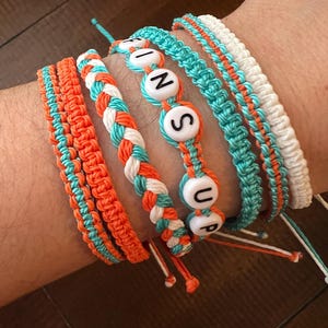 Op de afbeelding: Een set van zes vriendschapsarmbanden met oranje, wit en turquoise touw. De armbanden hebben witte kralen met de letters "FINS UP" erop.