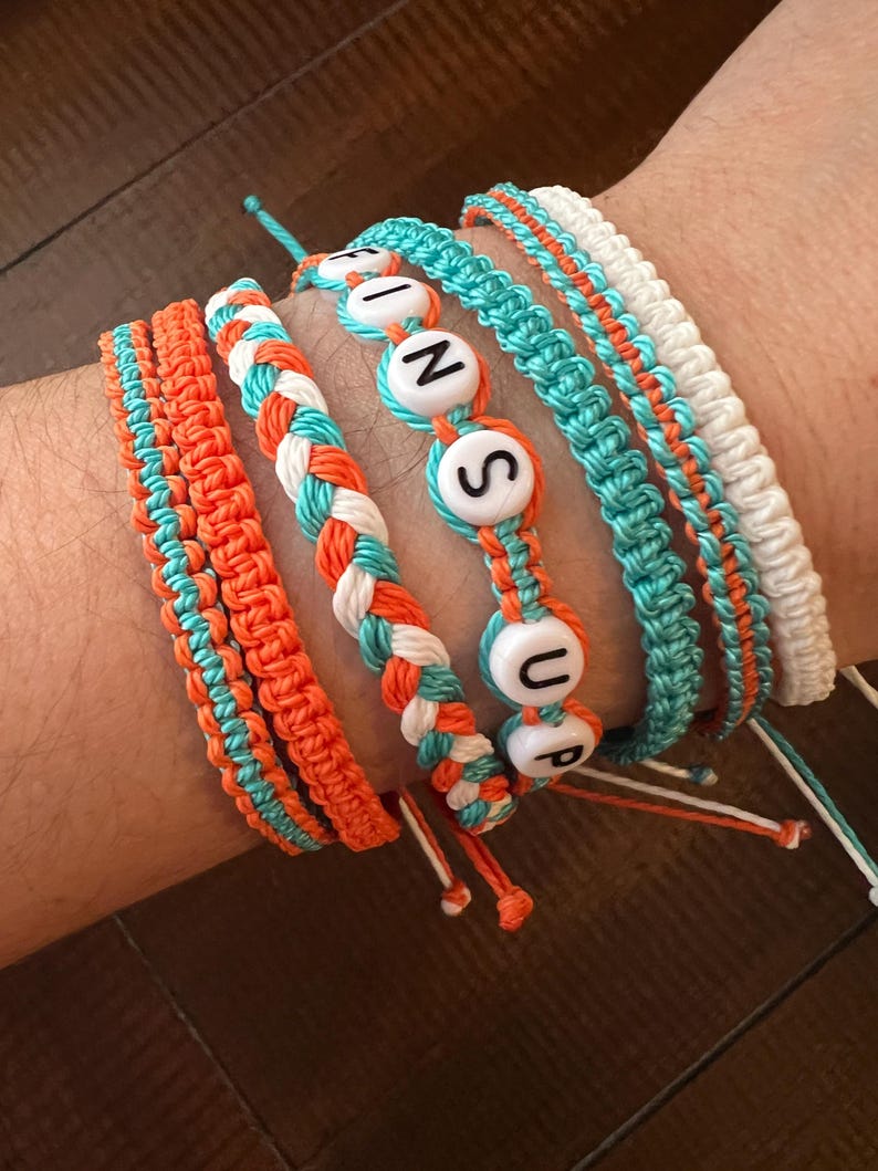 Op de afbeelding: Een set van zes vriendschapsarmbanden met oranje, wit en turquoise touw. De armbanden hebben witte kralen met de letters "FINS UP" erop.