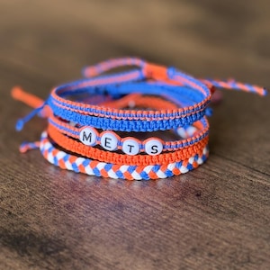 Op de afbeelding: Vier handgemaakte macrame armbanden in blauw, oranje en wit. Eén armband spelt "METS" met letterkralen. De armbanden zijn gestapeld op een houten oppervlak. Een fan accessoire.