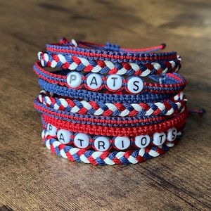 May include: Stack of handmade bracelets in red, white, and blue. The bracelets are braided with the word "PATS" and "PATRIOTS" spelled out in white letter beads. The bracelets are stacked on a wooden surface.