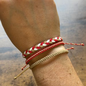 May include: Four handmade bracelets in red, white, and tan colors are displayed on a wrist. The top bracelet is braided with red, white, and tan threads. The other three bracelets are made with a woven design.