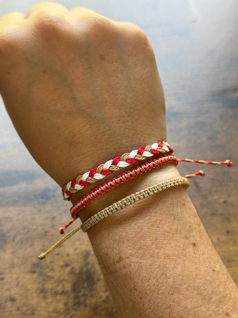 May include: Four handmade bracelets in red, white, and tan colors are displayed on a wrist. The top bracelet is braided with red, white, and tan threads. The other three bracelets are made with a woven design.