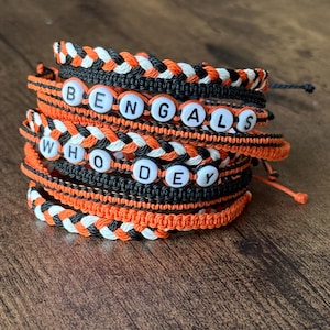 Könnte beinhalten: Stapel handgefertigter Armbänder in Orange, Schwarz und Weiß. Einige Armbänder buchstabieren "BENGALS" und "WHO DEY" mit weißen Buchstabenperlen. Die Armbänder bestehen aus geflochtenem und geknotetem Kordel und werden auf einer Holzoberfläche präsentiert.