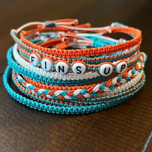 Op de afbeelding: Een set van zes kleurrijke vriendschapsarmbanden met het woord "FINS UP" gespeld in witte kralen. De armbanden zijn gemaakt van oranje, wit en turquoise touw.