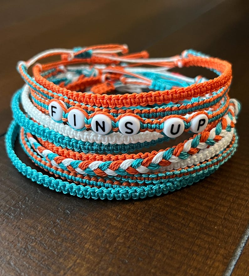 Op de afbeelding: Een set van zes kleurrijke vriendschapsarmbanden met het woord "FINS UP" gespeld in witte kralen. De armbanden zijn gemaakt van oranje, wit en turquoise touw.