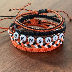 May include: A stack of three handmade cord bracelets in black, orange, and white. One bracelet features white round beads with black letters spelling BENGALS. The other two bracelets are braided with black and orange cord, featuring adjustable knots.