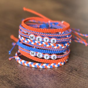 May include: Stack of handmade bracelets in orange, blue, and white. Some bracelets feature the word "METS" spelled out with white letter beads. The bracelets are made of braided and knotted cord.