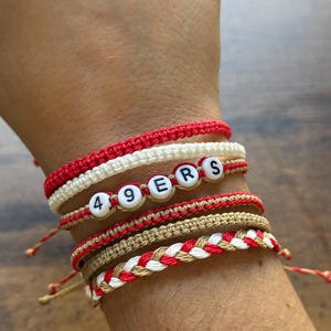 May include: A collection of handmade bracelets in red, white, and tan. One bracelet spells out "49ERS" with letter beads. The bracelets are made of woven and knotted cord, with adjustable ties.