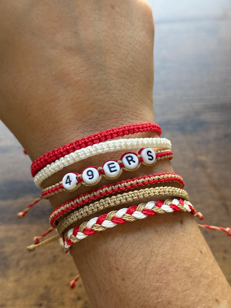 May include: A collection of handmade bracelets in red, white, and tan. One bracelet spells out "49ERS" with letter beads. The bracelets are made of woven and knotted cord, with adjustable ties.