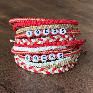 May include: Stack of handmade macrame bracelets in red, white, and tan. Each bracelet features the letters "49ERS" spelled out with white letter beads. The bracelets are stacked on a wooden surface.