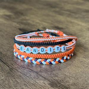 Può includere: Pila di braccialetti dell'amicizia fatti a mano in arancione, nero, bianco e viola. Un braccialetto riporta la scritta "BANDITS" con perline bianche. I braccialetti sono realizzati in cordoncino intrecciato e sono esposti su una superficie di legno.