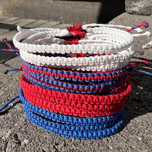 May include: A stack of colorful friendship bracelets in red, white, and blue. The bracelets are made of woven string and are tied together in a knot.