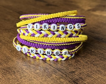 May include: Stack of handmade bracelets in purple, yellow, and white. The bracelets are braided and feature the word "VIKINGS" spelled out in white letter beads. The bracelets are stacked on a wooden surface.
