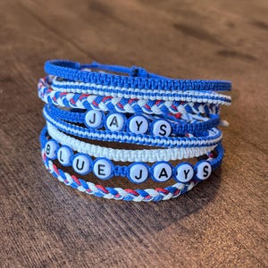 May include: A collection of blue, white, and red braided bracelets. Some bracelets feature white letter beads spelling "JAYS" and "BLUE JAYS". The bracelets are displayed on a wooden surface.