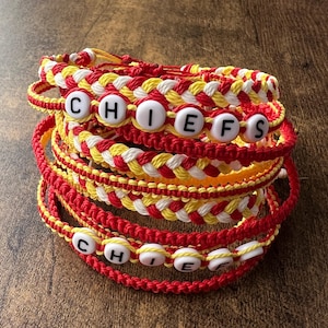 Könnte beinhalten: Ein Stapel handgefertigter Armbänder in Rot, Gelb und Weiß. Einige Armbänder buchstabieren das Wort "CHIEFS" mit weißen Buchstabenperlen. Die Armbänder bestehen aus geflochtenen und geknoteten Kordeln und werden auf einer Holzoberfläche präsentiert.