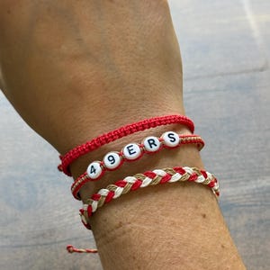 May include: A collection of handmade bracelets in red, white, and tan. One bracelet spells out "49ERS" with white letter beads. The other bracelets are braided or woven with red and white threads. The bracelets are worn on the wrist.