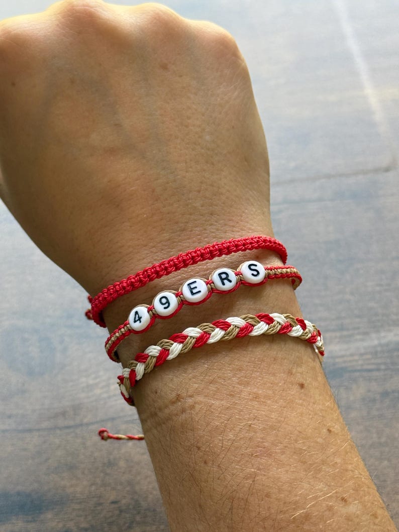 May include: A collection of handmade bracelets in red, white, and tan. One bracelet spells out "49ERS" with white letter beads. The other bracelets are braided or woven with red and white threads. The bracelets are worn on the wrist.