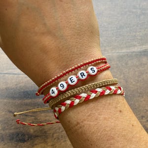 May include: A collection of handmade bracelets in red, white, and tan. One bracelet spells out "49ERS" with letter beads. Other bracelets feature braided and knotted designs. The bracelets are worn on a wrist.