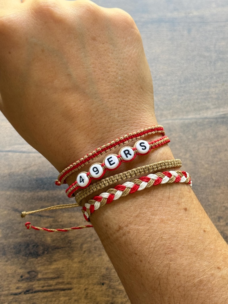 May include: A collection of handmade bracelets in red, white, and tan. One bracelet spells out "49ERS" with letter beads. Other bracelets feature braided and knotted designs. The bracelets are worn on a wrist.