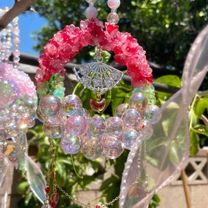 May include: A decorative suncatcher with a floral design. The suncatcher features a ring of pink flowers, iridescent bubble-like beads, and dangling charms. A silver fan-shaped charm and a red heart-shaped charm hang from the centre. The text "made by kool" is in the upper left corner.
