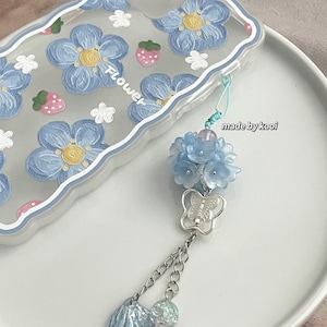 Op de afbeelding: Een transparante telefoonhoes met een ontwerp van blauwe bloemen en aardbeien. De hoes heeft een blauwe en witte kralenhanger met een vlinder en schelpaccenten. De hanger is gelabeld "made by kooi".