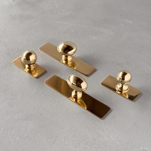 May include: Four gold cabinet pulls, each with a rectangular base and a rounded knob. The knobs have a shiny, reflective surface. The pulls are arranged on a light grey surface.