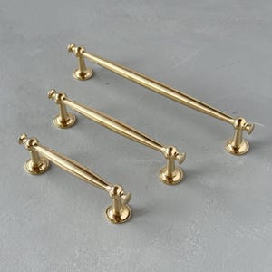 May include: Three gold-coloured cabinet pulls of varying sizes are arranged on a grey surface. Each pull features a rounded centre and decorative end pieces. The pulls are designed for easy gripping and add a touch of elegance.
