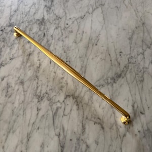 May include: A long, gold cabinet pull with a ridged centre and rounded ends. The hardware is shown against a marble background. This decorative pull is suitable for drawers and cabinets.
