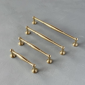 Può includere: Quattro maniglie dorate per mobili di diverse dimensioni sono disposte diagonalmente su una superficie grigia. Ogni maniglia ha un centro cilindrico e estremità arrotondate, con una base decorativa. Le maniglie sono in metallo e hanno una finitura lucida.