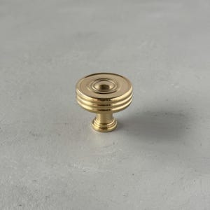 May include: A gold-coloured cabinet knob with a circular design. The knob features a flat top with concentric circles and three horizontal ridges below. The base is a smaller, rounded shape. The hardware is made of metal.