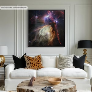 Peut inclure: Un canapé blanc avec des coussins de couleur noire, marron et blanche à motifs devant une impression encadrée en noir d'une nébuleuse colorée dans l'espace. L'impression est accrochée à un mur blanc avec des moulures blanches.