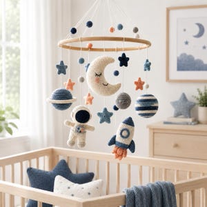 May include: A space-themed nursery mobile featuring a circular wooden frame with hanging crocheted elements including a smiling crescent moon, an astronaut, a rocket ship, planets with rings, and various stars in shades of blue, cream, and peach.