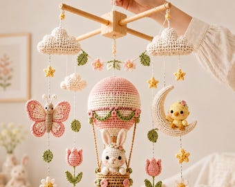 Könnte beinhalten: Ein gehäkeltes Mobile für das Kinderzimmer mit einem zentralen Heißluftballon, in dessen Korb ein weißer Hase sitzt, umgeben von hängenden Elementen wie einem rosa Schmetterling, einem gelben Küken auf einer Mondsichel, Wolken, Blumen und Blättern.