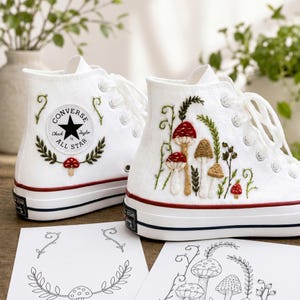 Op de afbeelding: Een paar witte hoge canvas sneakers met aangepast borduurwerk van rode en bruine paddenstoelen met groene ranken. De linkerschoen toont het ronde Converse All Star-logo, terwijl de rechterschoen een gedetailleerde botanische scène laat zien.