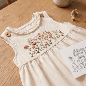 May include: An off-white sleeveless romper featuring a ruffled neckline and two wooden buttons at the shoulders. The chest is decorated with detailed floral embroidery in shades of orange, brown, and green, accompanied by a matching line art card.