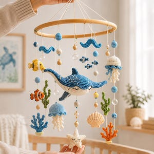 May include: A hanging nursery mobile featuring crocheted sea creatures including a blue stingray, a yellow fish, a clownfish, a starfish, and two jellyfish. The mobile includes green and orange coral, blue waves, and wooden beads on white strings.
