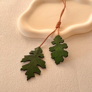 May include: A green leaf necklace with two leaves hanging from a brown cord. The leaves are made of wood and have a detailed vein pattern.