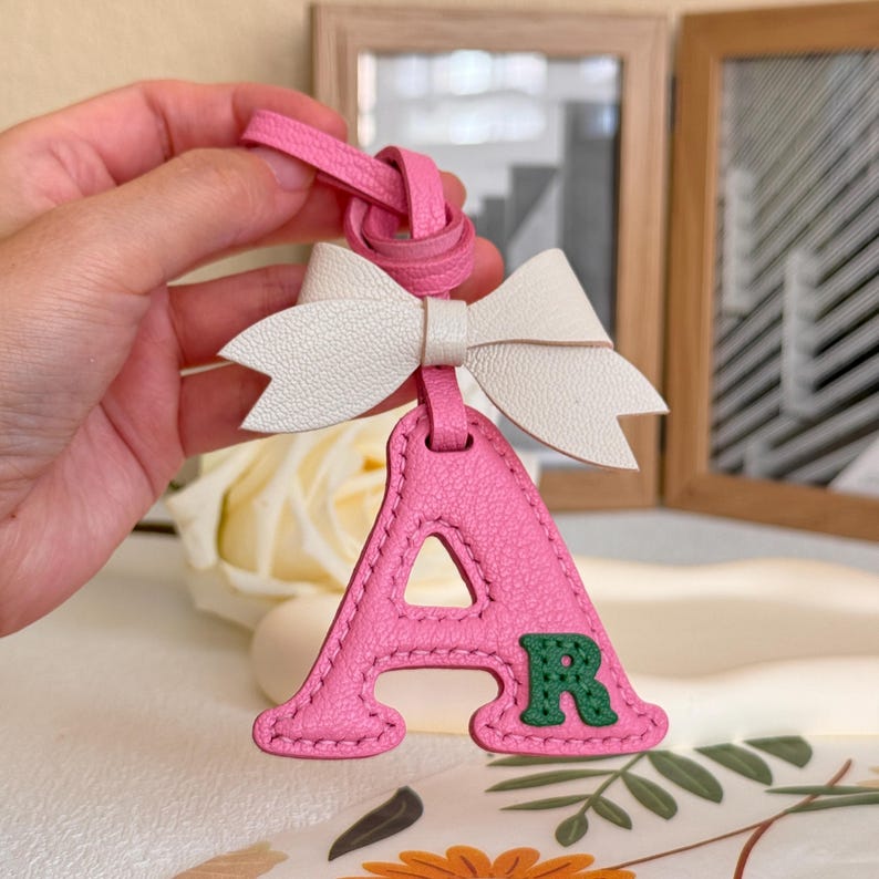 Puede incluir: Llavero de cuero rosa con un lazo blanco y una letra A rosa con una letra R verde en su interior. El llavero est&aacute; hecho de cuero y tiene un dise&ntilde;o cosido.