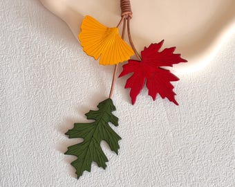 May include: A decorative hanging ornament featuring three distinct leaf shapes attached to a thin brown cord. The leaves include a bright yellow ginkgo leaf, a vibrant red maple leaf, and a dark forest green oak leaf, all with detailed veining.