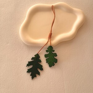 May include: Two green leather leaves attached to a brown leather cord. The leaves are shaped like oak leaves and have a textured surface.