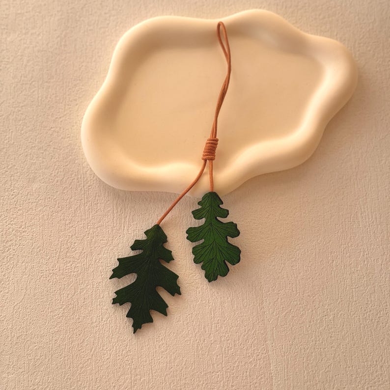 May include: Two green leather leaves attached to a brown leather cord. The leaves are shaped like oak leaves and have a textured surface.