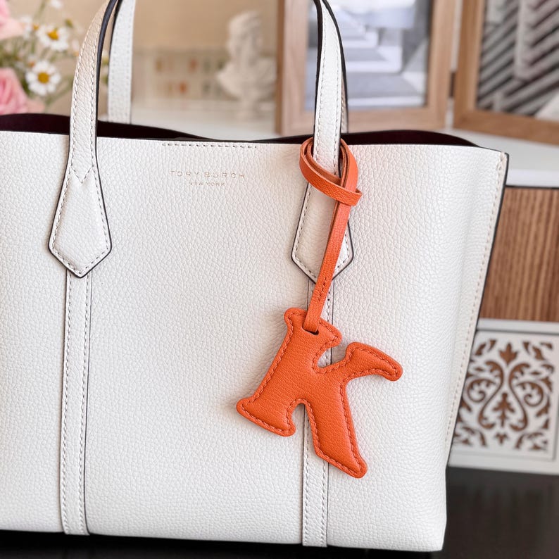 May include: White leather tote bag with a matching handle and a bright orange leather letter "K" charm. The bag has a minimalist design with the brand name "TORY BURCH" embossed on the front.