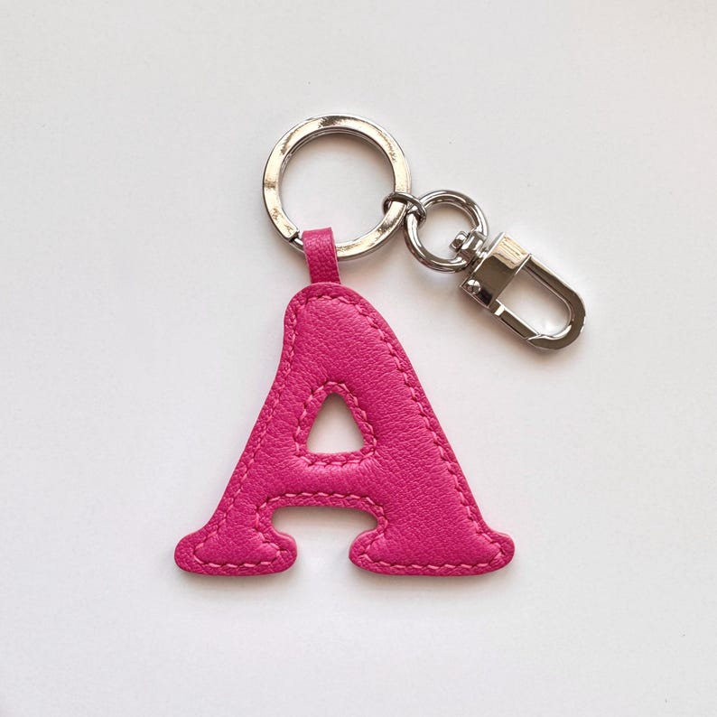 May include: A hot pink leather keychain featuring the letter "A". The letter is stitched with a matching pink thread and attached to a silver-tone keyring and clasp. The keychain is a personalized accessory.