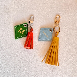 May include: Two leather keychains with tassels. One keychain is green with the letter "M" and a red tassel. The other keychain is blue with the word "Isabella" and a yellow tassel.