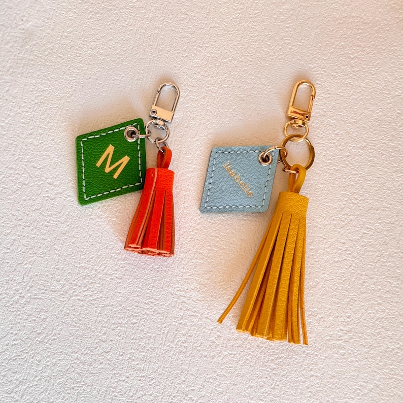May include: Two leather keychains with tassels. One keychain is green with the letter "M" and a red tassel. The other keychain is blue with the word "Isabella" and a yellow tassel.