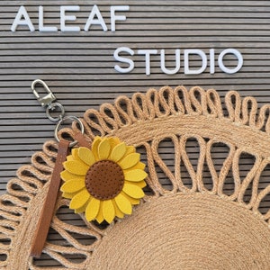 May include: A yellow sunflower keychain with a brown center and a brown leather strap attached to a silver key ring. The keychain is on a brown woven mat with the words "ALEAF STUDIO" in white letters on a gray background.